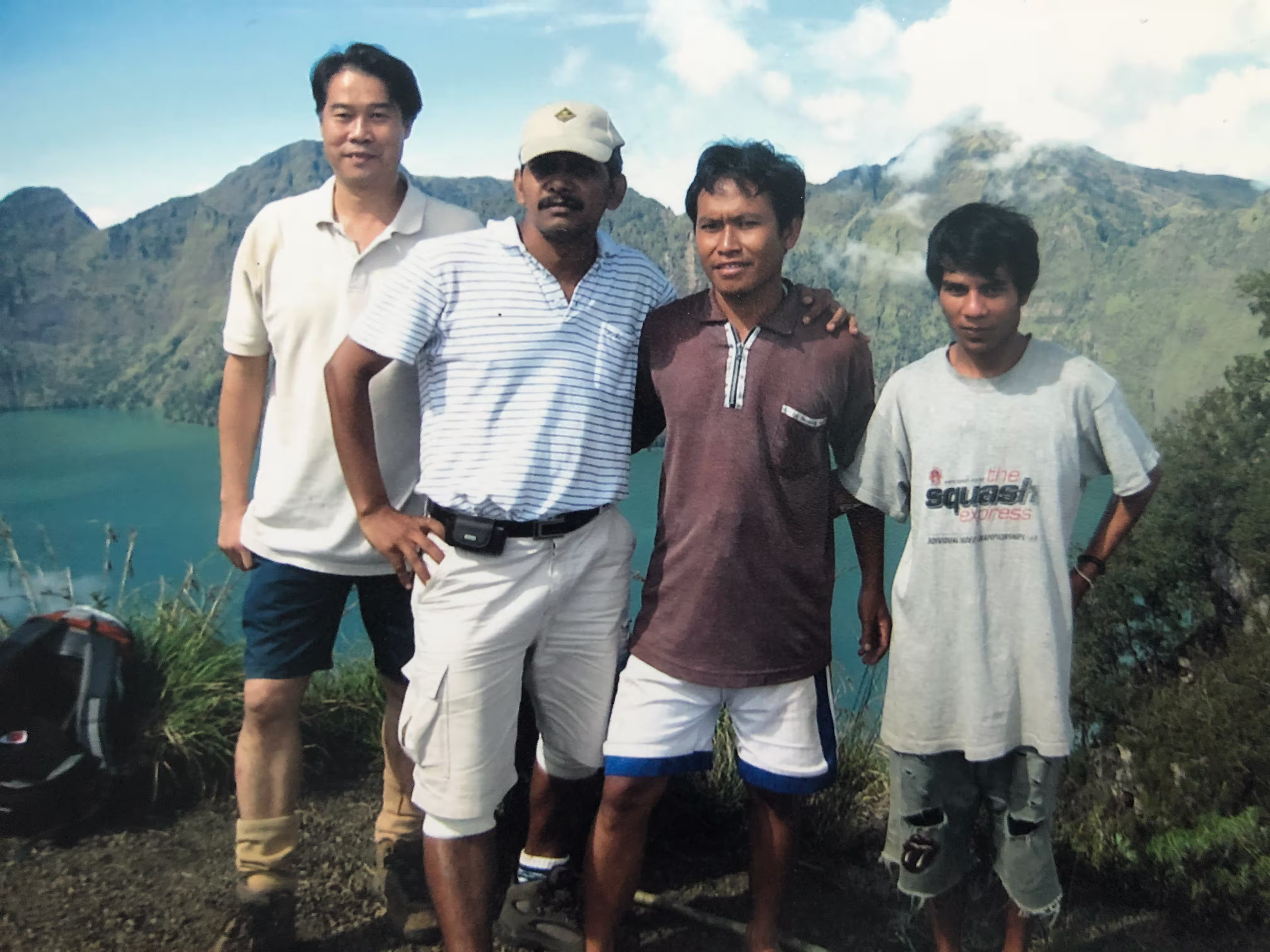 Four local guides and hikers posing together on the crater rim overlooking the turquoise Segara Anak lake on a trek organized by go2rinjani