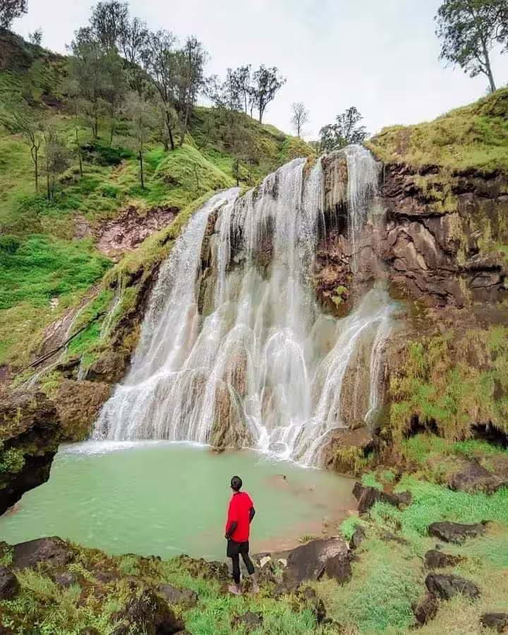 Visitor watching a wide waterfall cascade over mossy rocks into an emerald pool on the slopes of Mount Rinjani
