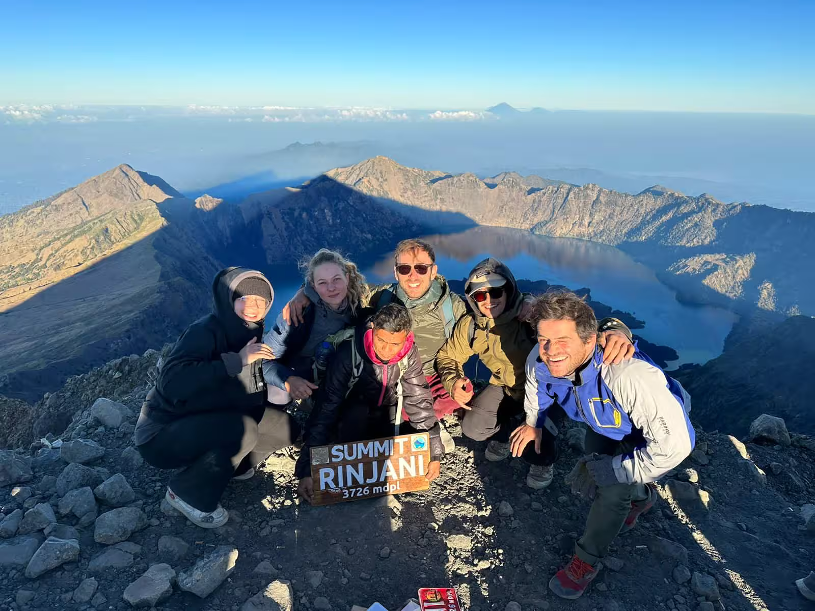 Group of trekkers and their guide posing with the Summit Rinjani 3,726m sign above the crater lake after completing a Mount Rinjani summit trek 2 days via Sembalun