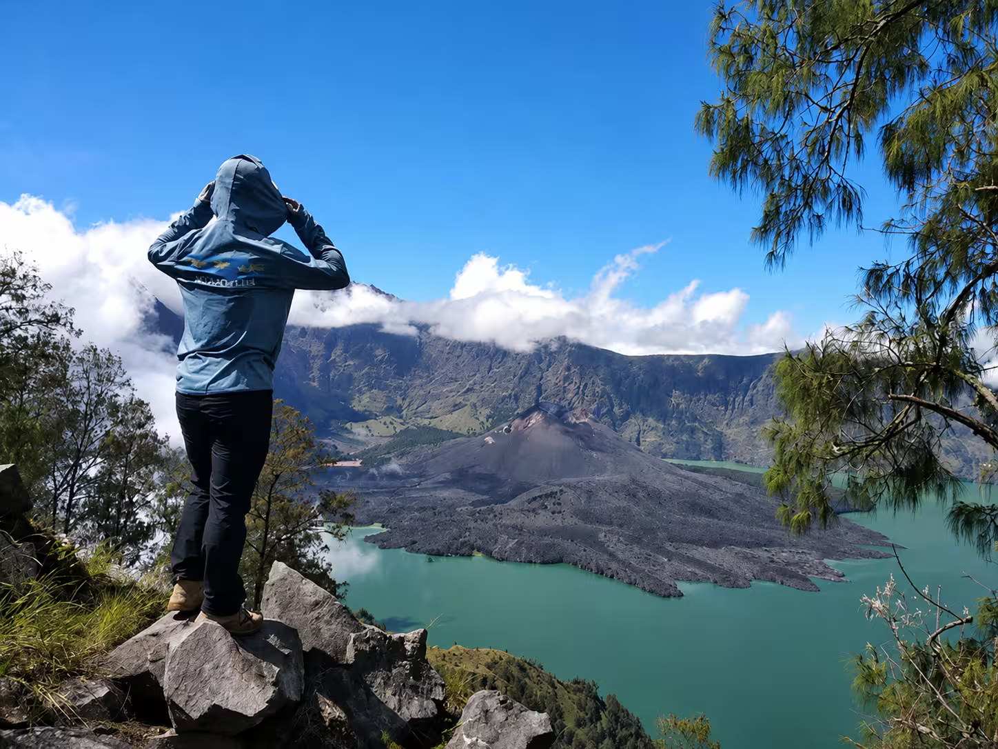Trekker standing on a rocky outcrop at the Senaru crater rim gazing at the turquoise Segara Anak lake and Barujari volcano cone — the viewpoint reached on a Mount Rinjani 2-day Senaru crater rim trek