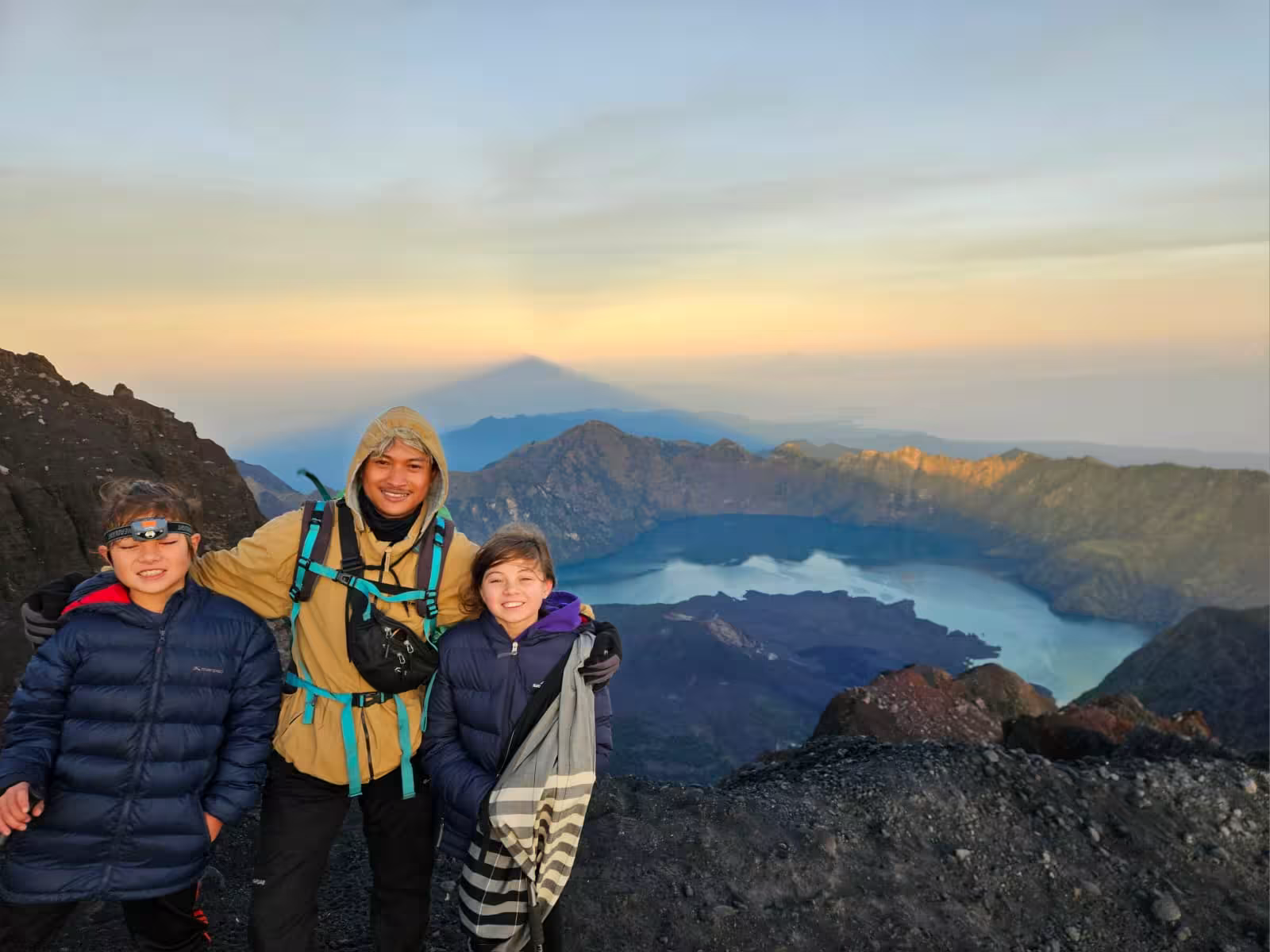 Guide and two young trekkers posing near the Rinjani summit at dawn with the mountain's shadow cast across the sky and Segara Anak lake below — a highlight of the Mount Rinjani 3-day Sembalun trek
