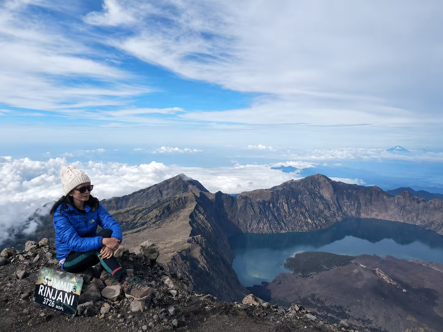 Woman sitting at the Rinjani 3,726m summit beside the summit sign with the crater rim, Segara Anak lake, and clouds stretching below — a rewarding moment on a Mount Rinjani 3-day Senaru trek