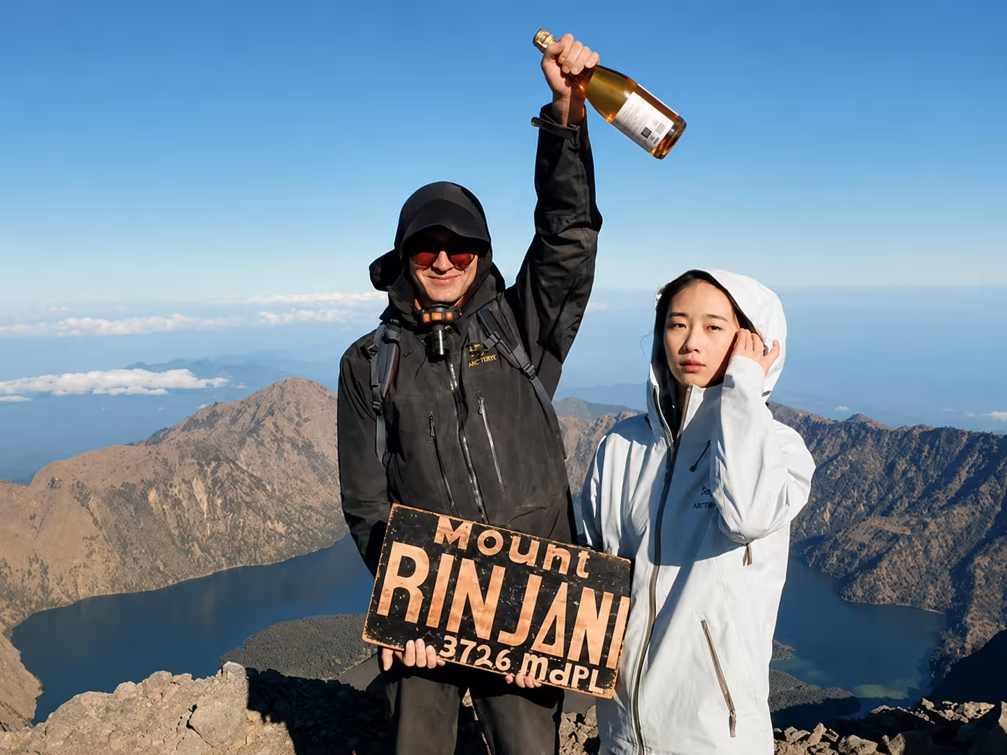 Couple celebrating at the Mount Rinjani 3,726m summit with a champagne bottle and the iconic summit sign, crater lake and caldera stretching behind them on a Mount Rinjani 4-day Sembalun expedition