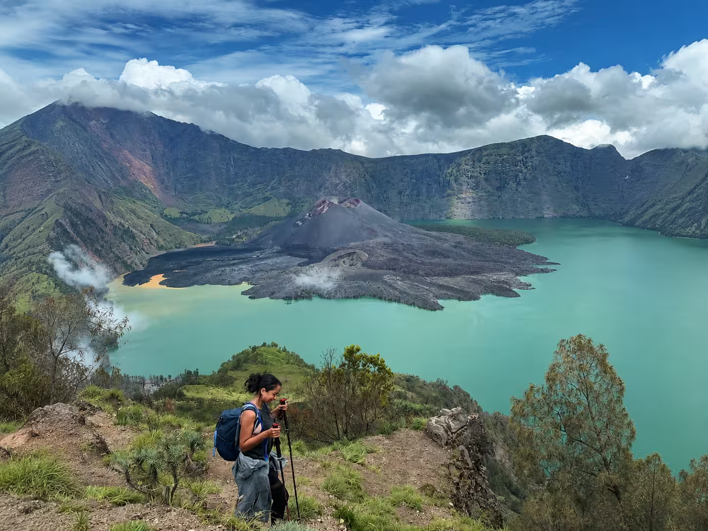 Trekker with backpack and poles on a grassy ridge overlooking the full Segara Anak lake, Barujari cone, and steaming hot springs during a Mount Rinjani 4-day Senaru trek