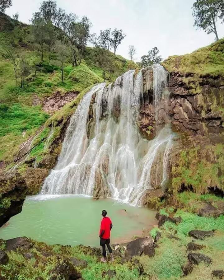 Visitor watching a wide waterfall cascade over mossy rocks into an emerald pool on the slopes of Mount Rinjani