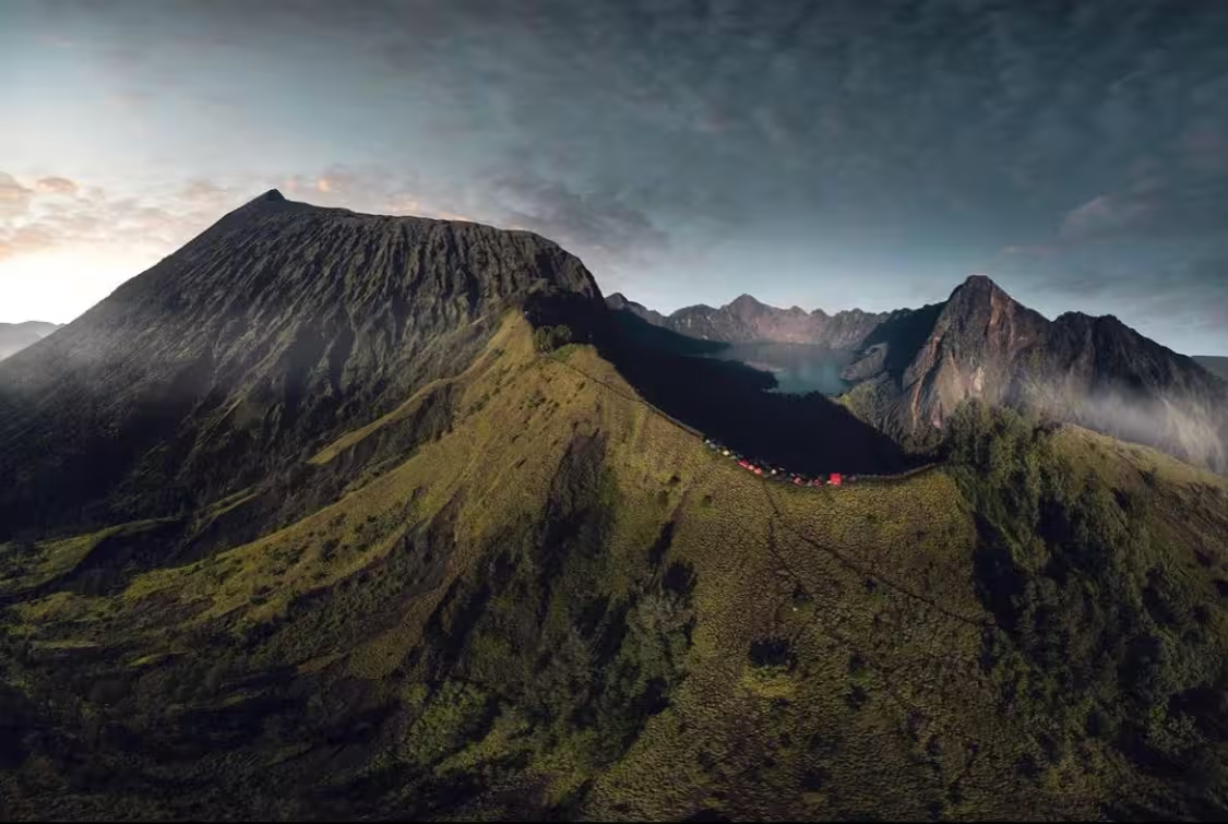 Panoramic drone shot of the Mount Rinjani trek showing the full crater rim, summit cone, and campsite nestled on the ridge at dusk