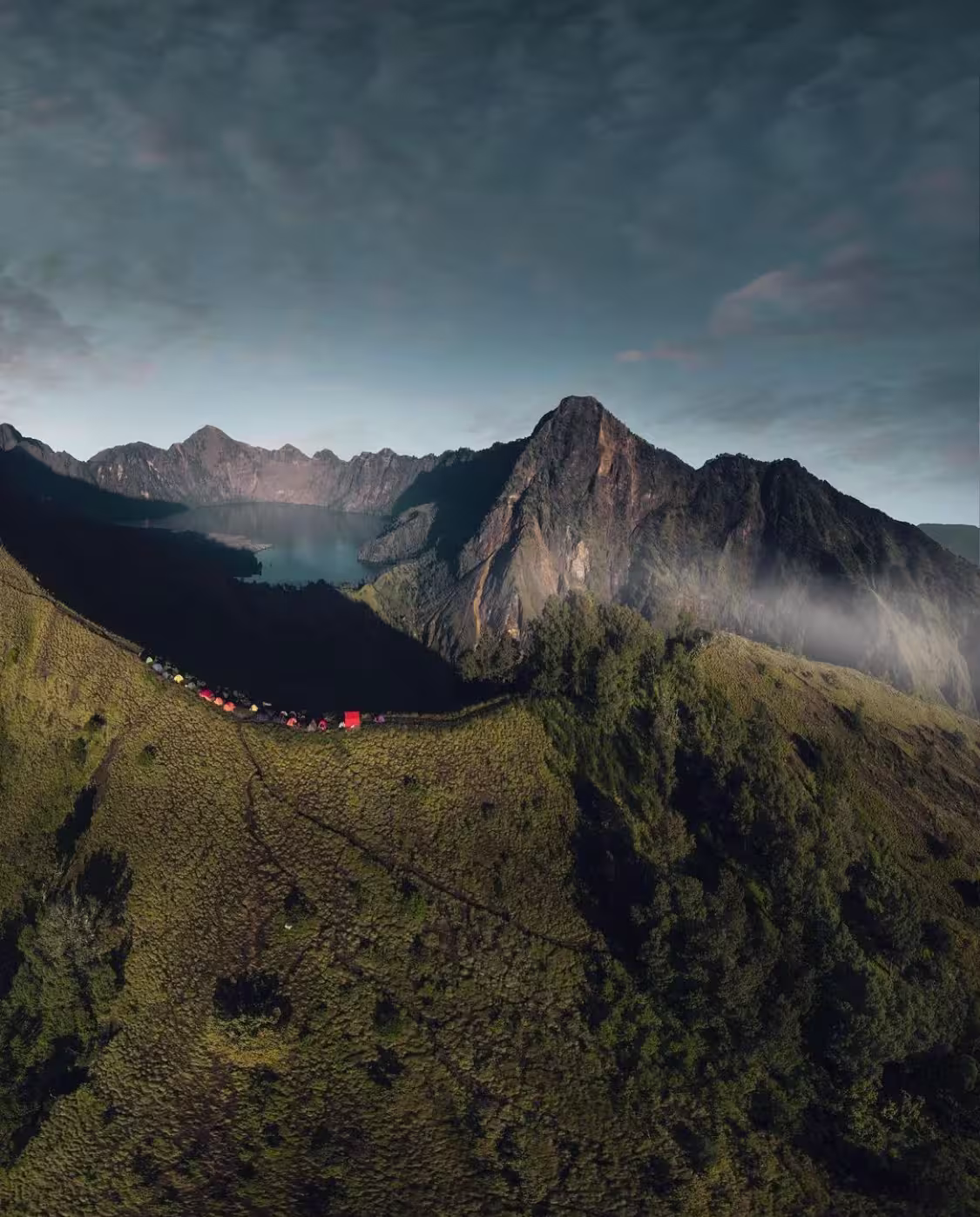 Aerial view of colorful camping tents on the green Sembalun crater rim with Segara Anak lake and Rinjani's rocky peaks behind