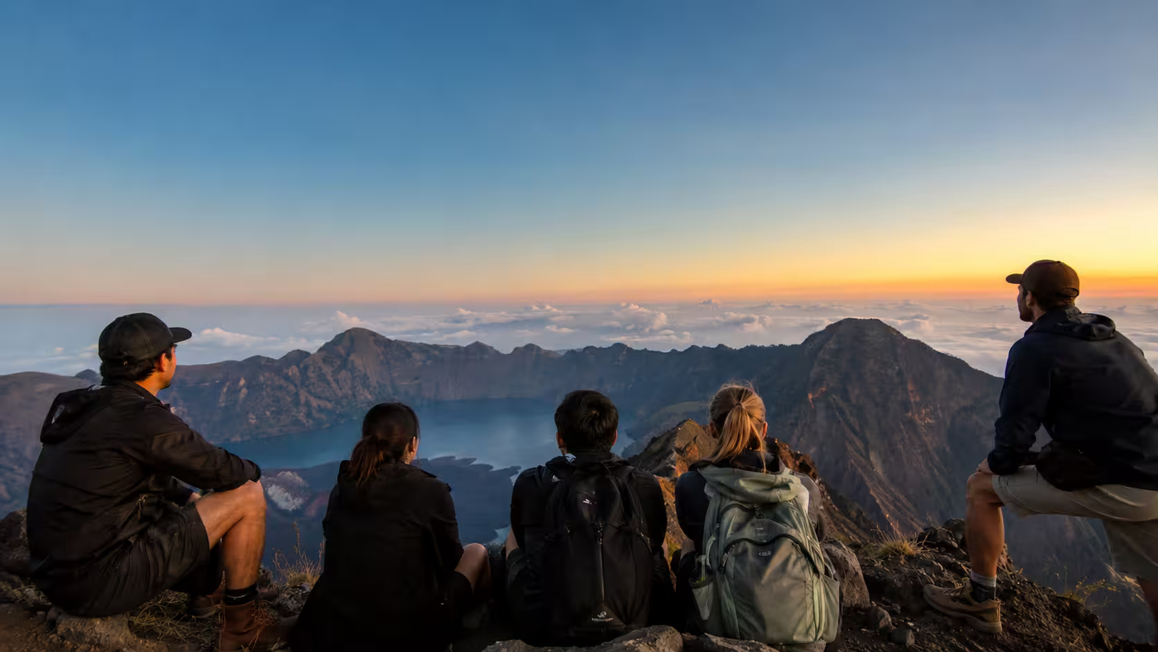 A group of adventurers sitting on the rocky edge of Mount Rinjani at sunrise, watching the morning light illuminate the clouds and crater lake.