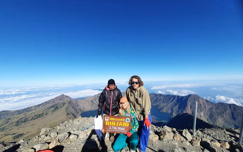 Three hikers stand triumphantly on Mount Rinjani's summit, posing beside a sign marking their trekking achievement Three hikers stand triumphantly on Mount Rinjani's summit, posing beside a sign marking their trekking achievement