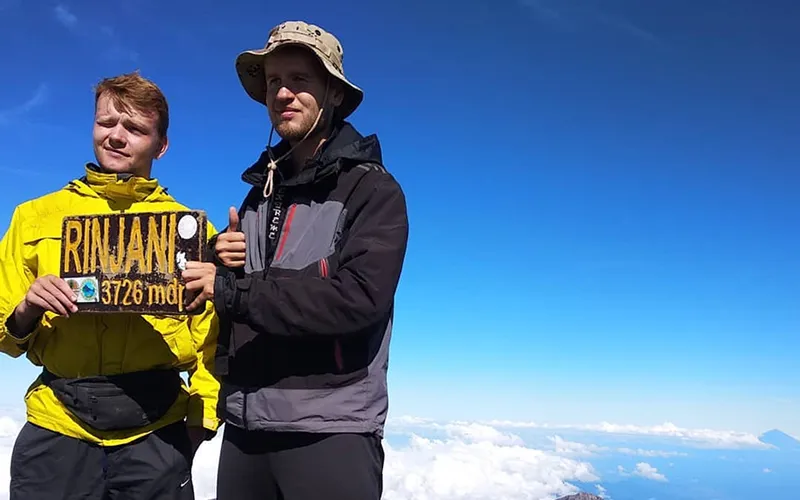 Two hikers at the top of Mount Rinjani, proudly displaying a sign after their three-day trek Two hikers at the top of Mount Rinjani, proudly displaying a sign after their three-day trek