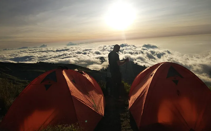 A hiker next to two tents on a mountain, capturing the adventure of the Mount Rinjani summit trek A hiker next to two tents on a mountain, capturing the adventure of the Mount Rinjani summit trek