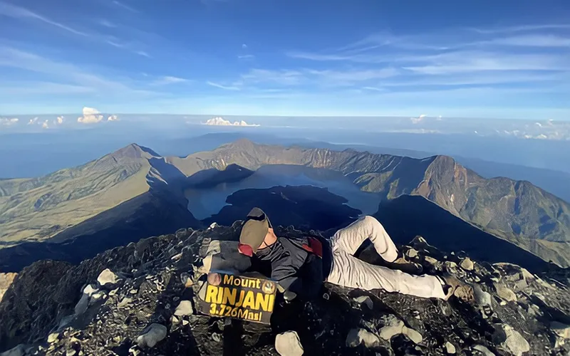 A man sits atop Mount Rinjani, holding a sign, enjoying the view after a hike on the Sembalun route A man sits atop Mount Rinjani, holding a sign, enjoying the view after a hike on the Sembalun route