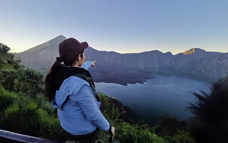 A woman enjoys the view of a peaceful lake and towering mountains, a moment from her Mount Rinjani hike A woman enjoys the view of a peaceful lake and towering mountains, a moment from her Mount Rinjani hike