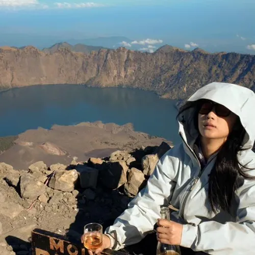 A woman in a rain jacket sits on a bench, enjoying a beer after hiking Mount Rinjani via the Sembalun route A woman in a rain jacket sits on a bench, enjoying a beer after hiking Mount Rinjani via the Sembalun route