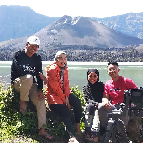 Friends sit on a rock near a serene lake, enjoying a break during their Mount Rinjani hike Friends sit on a rock near a serene lake, enjoying a break during their Mount Rinjani hike