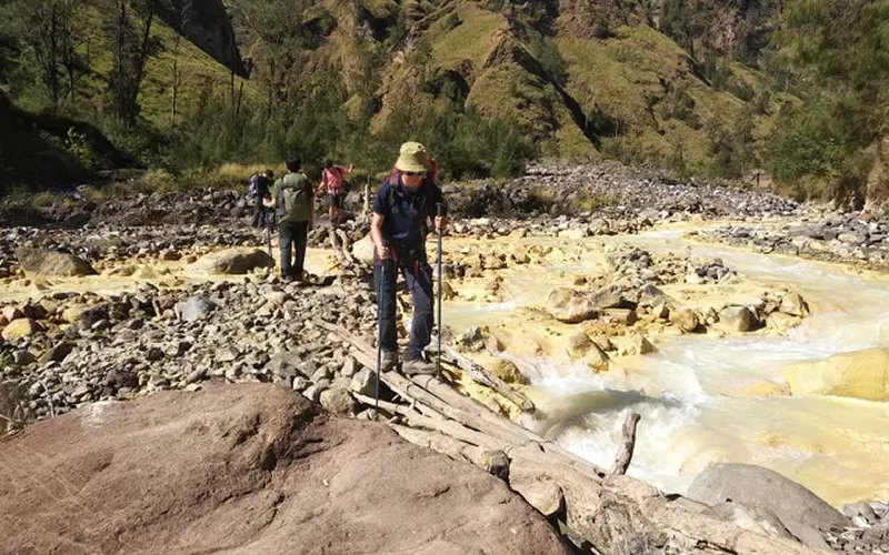 Hikers making their way across a rocky river, surrounded by debris, on the Mount Rinjani trek route via torean Hikers making their way across a rocky river, surrounded by debris, on the Mount Rinjani trek route via torean