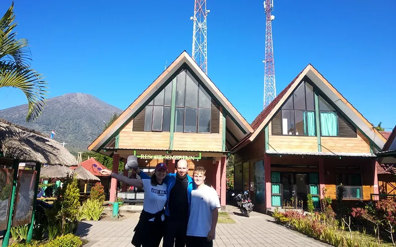 Three adventurers in front of a house, with a mountain looming behind, reflecting on their Mount Rinjani hike Three adventurers in front of a house, with a mountain looming behind, reflecting on their Mount Rinjani hike