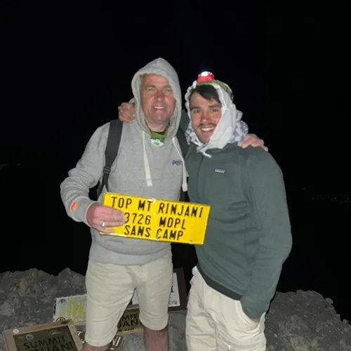 Two men on a mountain summit hold a sign celebrating their hike on the Sembalun route to Mount Rinjani Two men on a mountain summit hold a sign celebrating their hike on the Sembalun route to Mount Rinjani