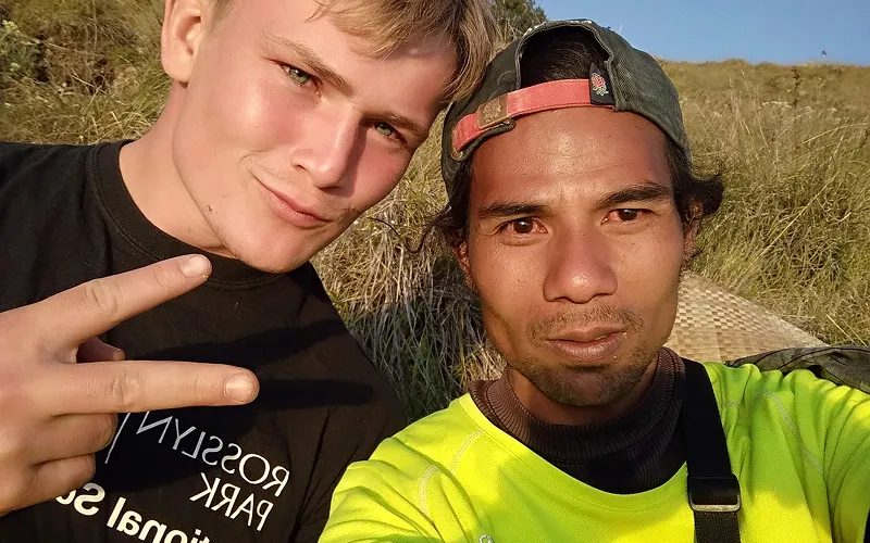 Two young men smiling and taking a selfie while sitting on the grass during their Mount Rinjani hike adventure Two young men smiling and taking a selfie while sitting on the grass during their Mount Rinjani hike adventure