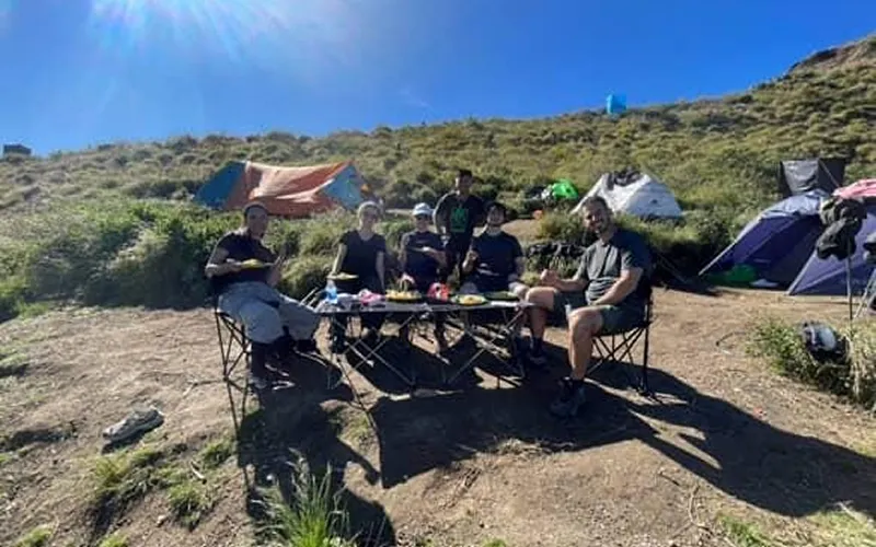 A group of hikers sitting at a table in the mountains, enjoying a meal during their Mount Rinjani trek on the Sembalun route A group of hikers sitting at a table in the mountains, enjoying a meal during their Mount Rinjani trek on the Sembalun route